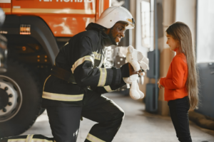 A Man Wearing Safety Gear Handing a Teddy Bear to a Girl Standing in front of Her